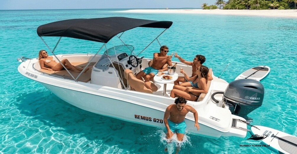 A white motorboat with a black canopy on a turquoise sea, a person in swim trunks, and a wakeboard. 1