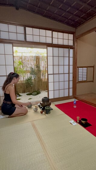 A person sitting on tatami mat in a traditional Japanese room with shoji screens, a tea ceremony setup, and a bamboo fence visible through the window. 3
