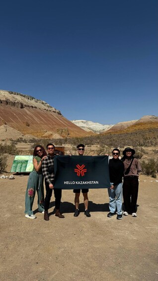 A group of people holding a flag with 'HELLO KAZAKHSTAN' text, standing in a desert landscape with mountains in the background. 1