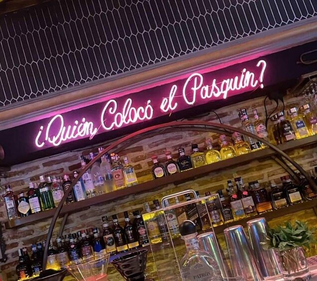 A bar with a neon sign reading '¿Quién Colocó el Pasquin?' and shelves stocked with various liquor bottles. 5