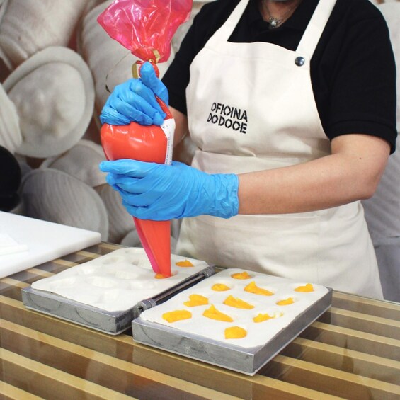 A person in a white apron and blue gloves using a red piping bag to fill a white tray with yellow substance. 1