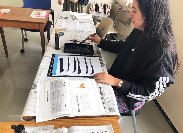 A person practicing calligraphy with a brush, surrounded by papers and a book on a table. 5