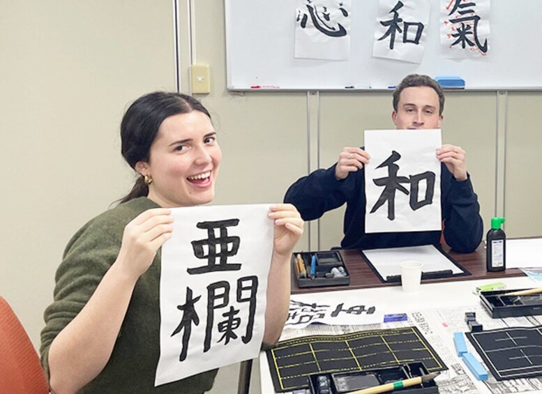 Two individuals holding papers with Japanese characters, a whiteboard with Chinese characters, and a table with stationery items. 1