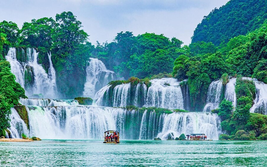A multi-tiered waterfall surrounded by lush greenery, with a boat tour visible in the foreground. 2