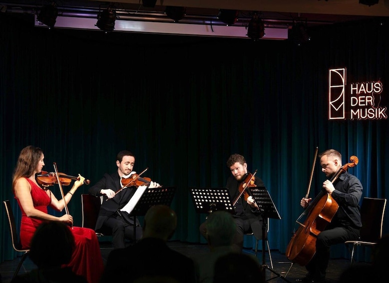 En stråkkvartett spelar på en scen med en mörk, draperad bakgrund och en logotyp med texten ”HAUS DER MUSIK” i det övre högra hörnet. 1