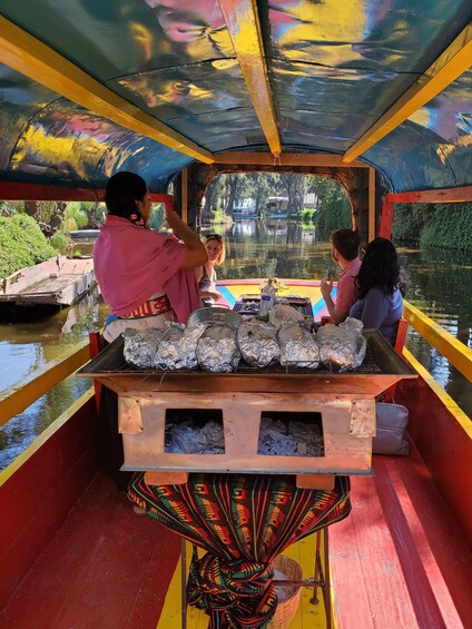 A colorful boat with people on board, a grill with foil-wrapped items, and a river with greenery and buildings in the background. 2