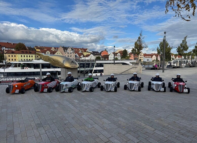 A group of people in go-karts on a paved area with buildings and a boat in the background. 3
