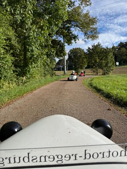 A dirt path with a white vehicle in the foreground and two people in red vehicles in the distance. 1