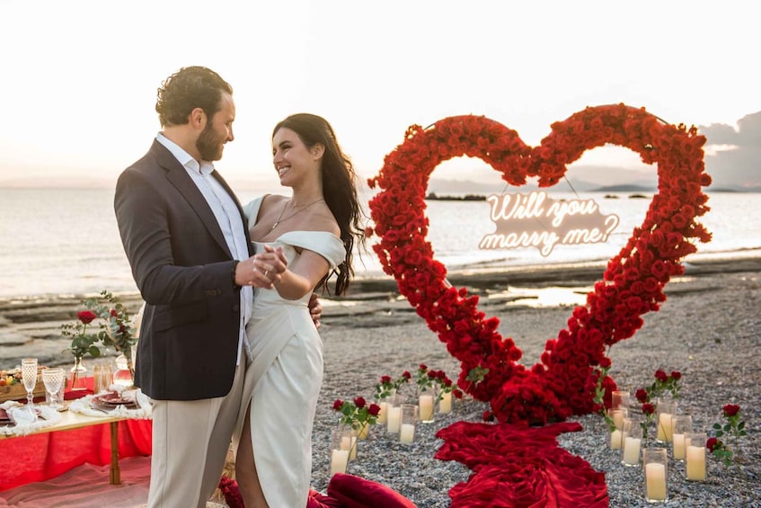 A couple standing by a heart-shaped floral arrangement with candles on a beach at sunset. 3