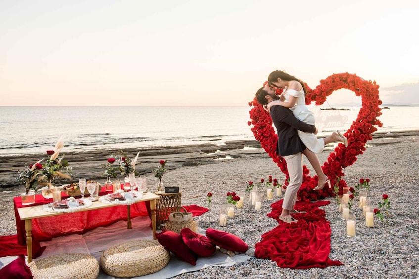 A beach setting with a heart-shaped floral arch, a table with red tablecloth, and candles. 1