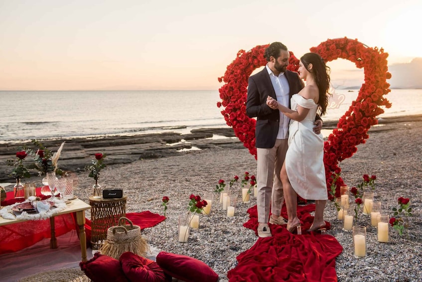 A couple standing in a heart-shaped floral arch on a beach with a table set for a romantic meal. 4