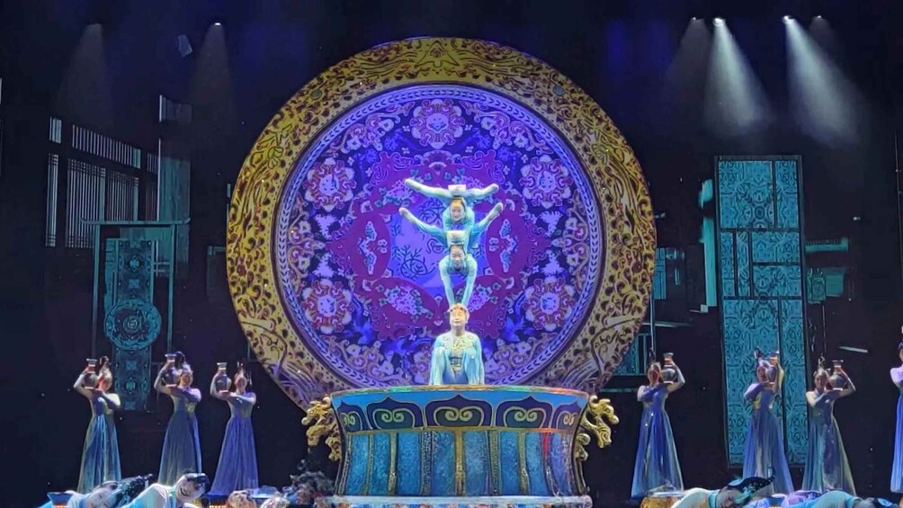 A stage with a central figure, surrounded by performers in blue dresses, against a backdrop of ornate patterns and architectural elements. 5