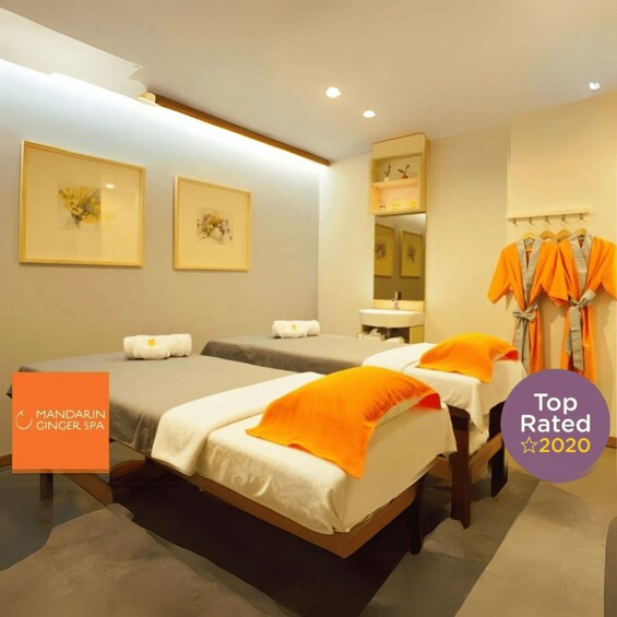 A spa room with two beds, orange pillows, a mirror, and a rack of robes. 1