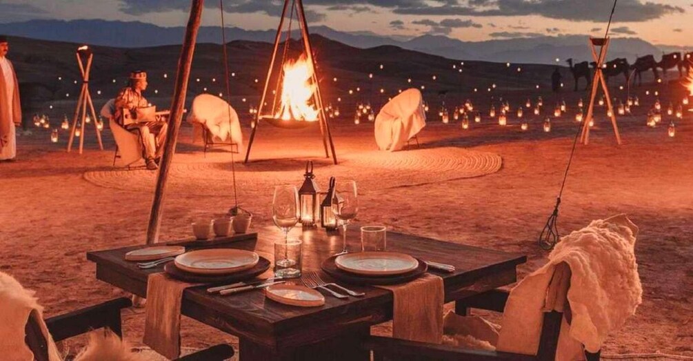 A desert campsite with a wooden table set for a meal, surrounded by tents and a campfire. 5