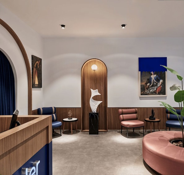A modern interior with a wooden reception desk, blue and pink seating, a white abstract sculpture, and two paintings on the walls. 5