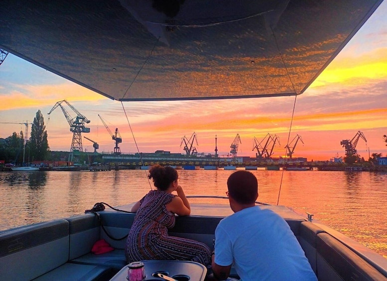 A boat with two people, a sunset, and industrial cranes in the background. 1