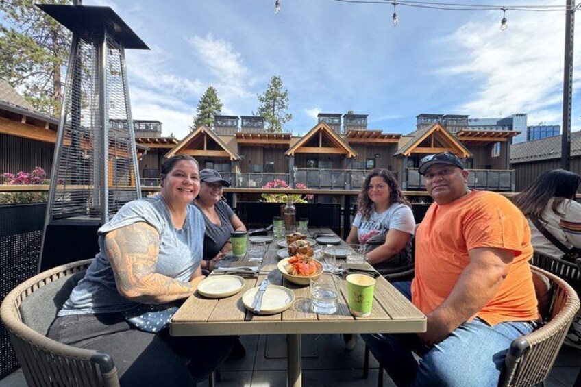 A group of people seated at an outdoor table with food and drinks, surrounded by wooden cabins. 1