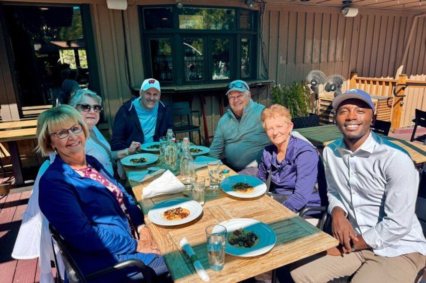 A group of people sitting at an outdoor table with plates of food, a fan, and a wooden deck. 3