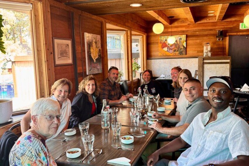 A group of people seated around a wooden table in a restaurant with wooden walls and ceiling, with various dishes and glasses on the table. 4