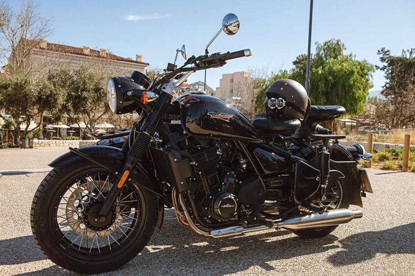 A black motorcycle parked on a gravel surface with trees and buildings in the background. 2