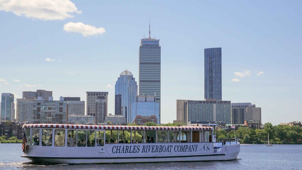 A riverboat with 'Charles Riverboat Company' written on its side, sailing on a river with a city skyline in the background. 1
