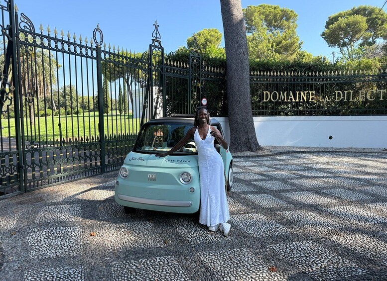 A woman in a white dress standing next to a vintage Fiat car in front of a gated entrance with a sign reading 'DOMAINE D'ELLEC.' 2