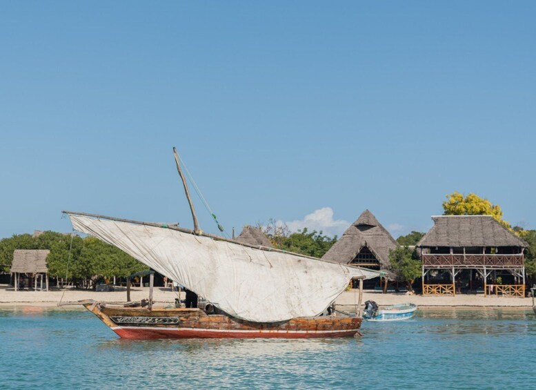 A traditional wooden boat with a sail on calm water, surrounded by thatched-roof buildings and a beach. 3