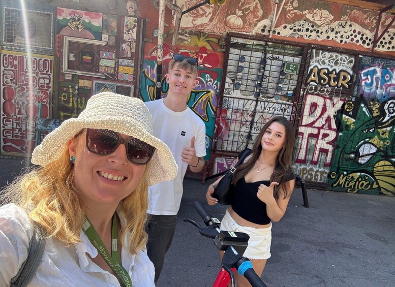 Una persona con un cappello bianco e occhiali da sole, un'altra con una camicia bianca e una terza con una maglietta nera e pantaloncini bianchi, in piedi di fronte a un muro ricoperto di graffiti. 1