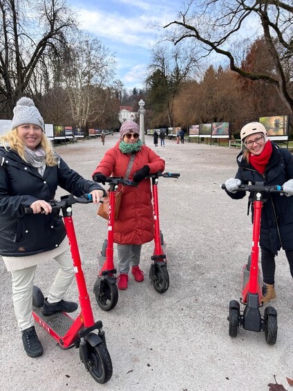 Three people riding red electric scooters in a park with bare trees and informational signs in the background. 2