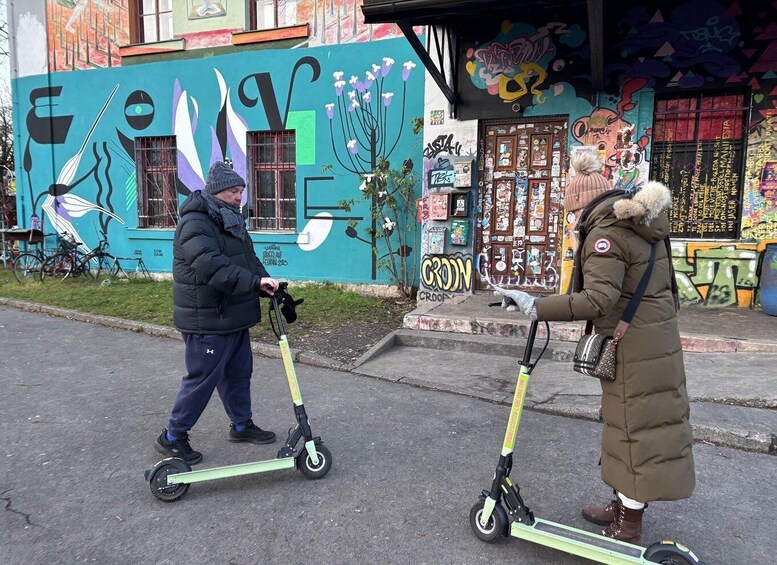Two people on electric scooters on a street with colorful graffiti on the buildings. 3