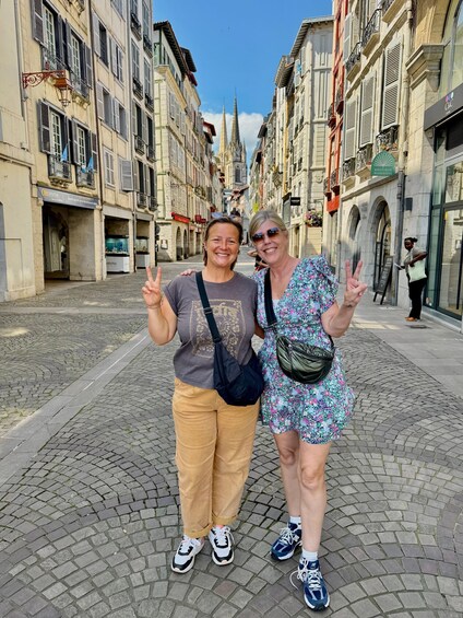 Two people posing on a cobblestone street with historic buildings and a church spire in the background. 2