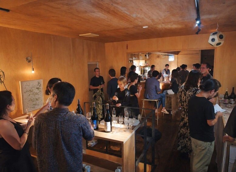 A group of people in a wooden interior space, some seated at tables with wine bottles and glasses, others standing or walking. 3