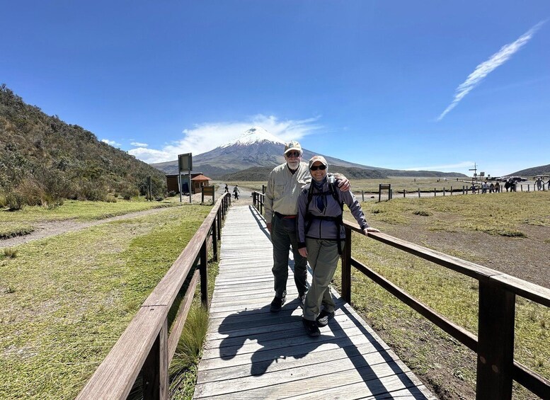 A wooden boardwalk with two people walking, a mountain in the background, and a small building to the left. 2