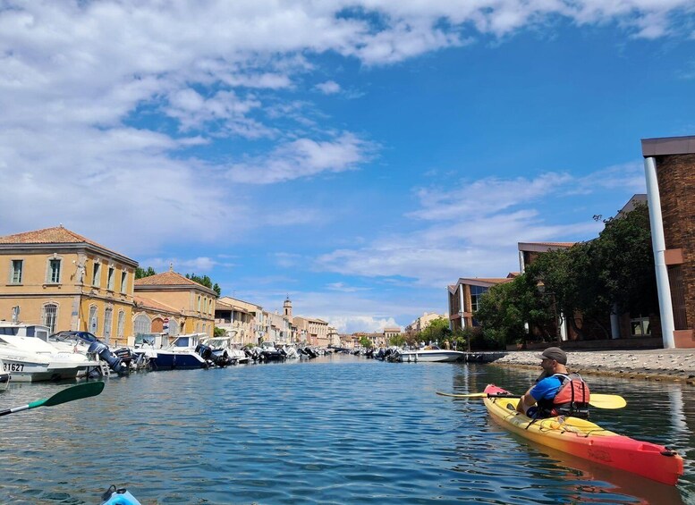 A person kayaking on a calm river, surrounded by historic buildings and parked boats. 5