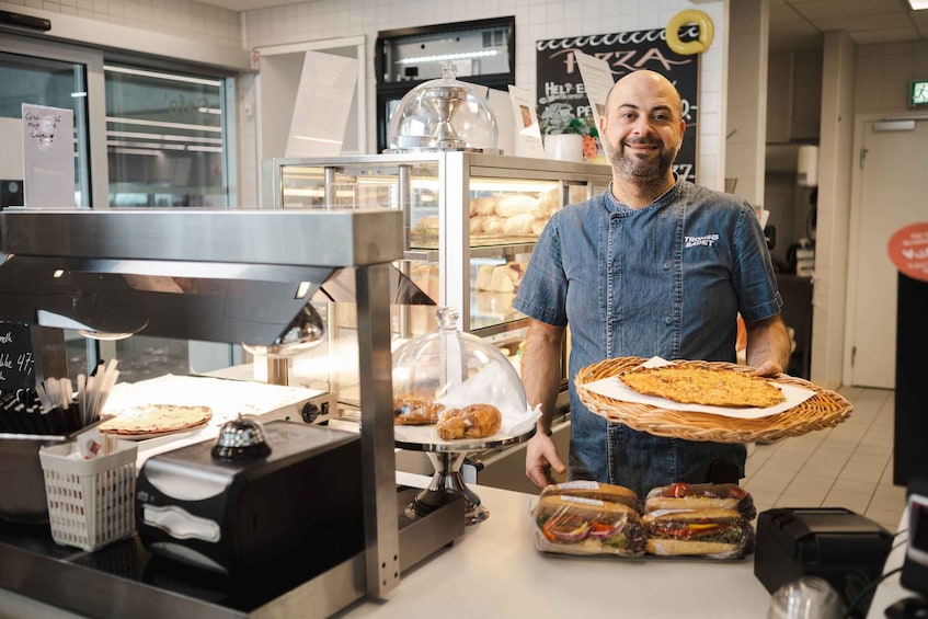 A person in a denim shirt holding a basket with a flatbread, standing in a bakery with bread display cases and a coffee machine. 3