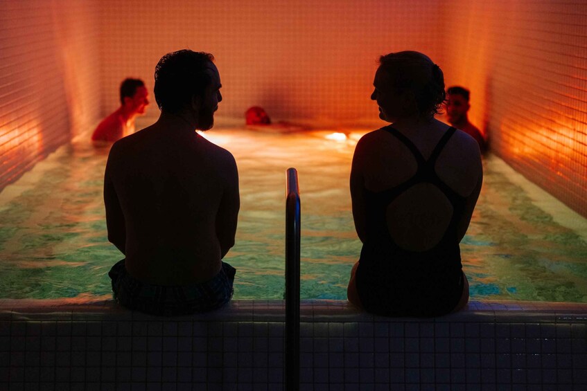Two people sitting by a pool with a tiled wall and a person in the water. 2