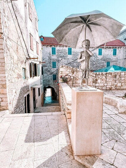 A narrow street with stone buildings, a statue, and a large umbrella. 1