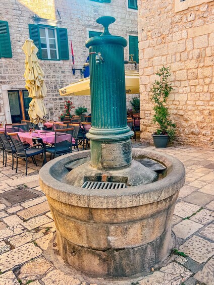 A traditional stone fountain with a green spout, surrounded by outdoor seating with checkered tablecloths and umbrellas. 5