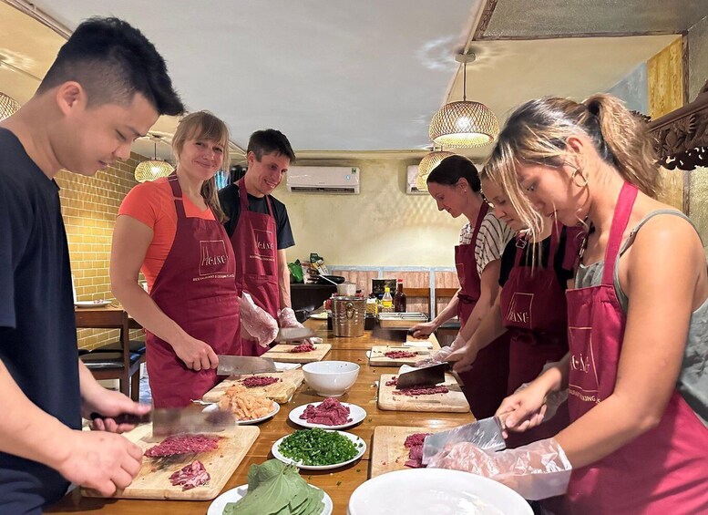 A group of people in a kitchen preparing food, wearing aprons, with various ingredients and utensils on the counter. 3