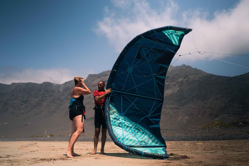 Two people with a large blue kite on a beach with mountains in the background. 1