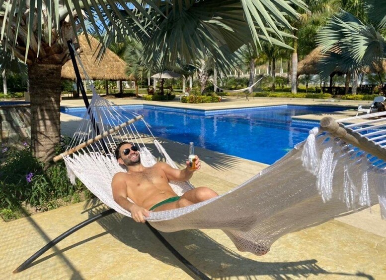A person relaxing in a hammock by a pool with palm trees and a thatched-roof structure in the background. 2