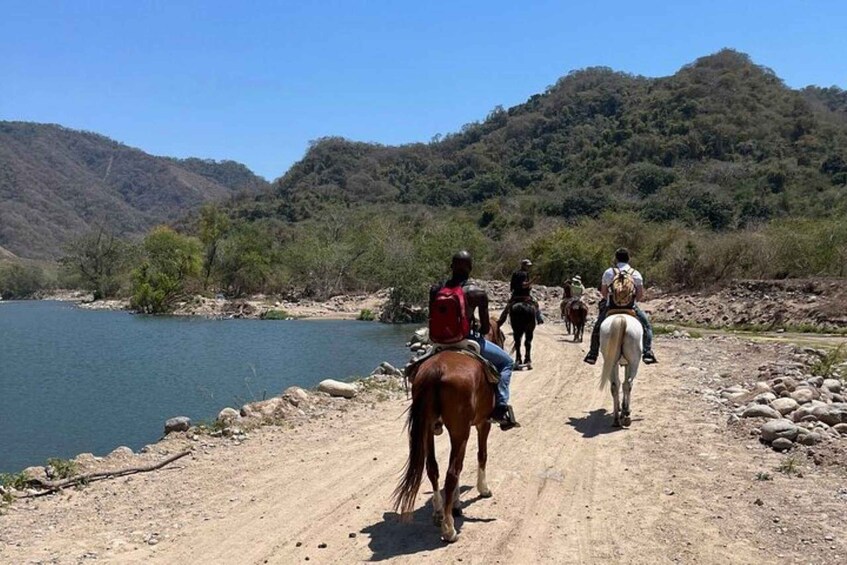 A group of people horseback riding along a dirt path beside a body of water, with hills in the background. 4