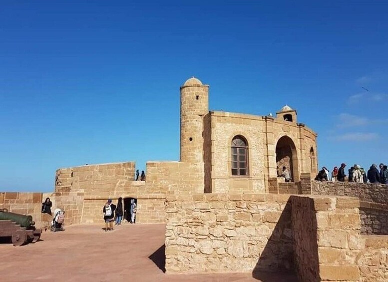 A historical stone building with arched windows and a tower, surrounded by a stone wall and a clear blue sky. 5
