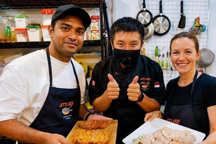 Three people in a kitchen, one holding a cutting board with food, another giving a thumbs up, and a third holding a plate of food. 2