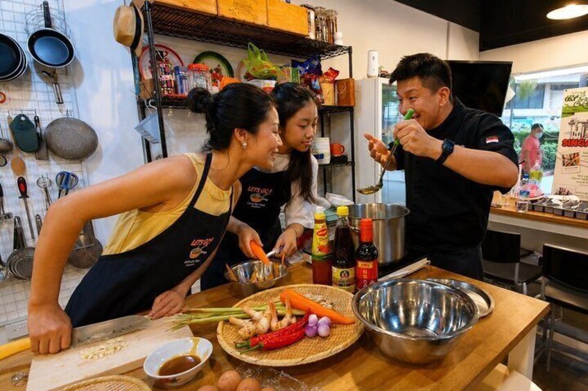 A kitchen with people preparing food, a cutting board, vegetables, and cooking utensils. 5