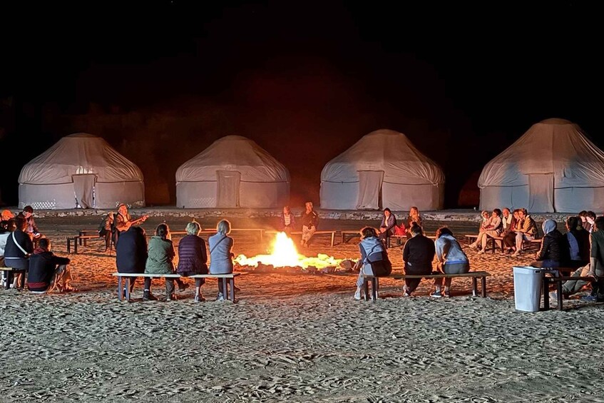 Yurts, a campfire, and people sitting around the fire on a sandy ground. 4