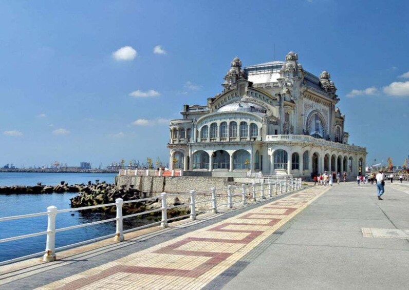 A grand, ornate building with arched windows and a domed roof, situated by a waterfront with a paved walkway and a railing. 3