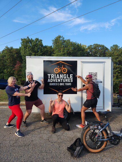 Four people posing in front of a sign for Triangle Adventures guided e-bike tours. 3