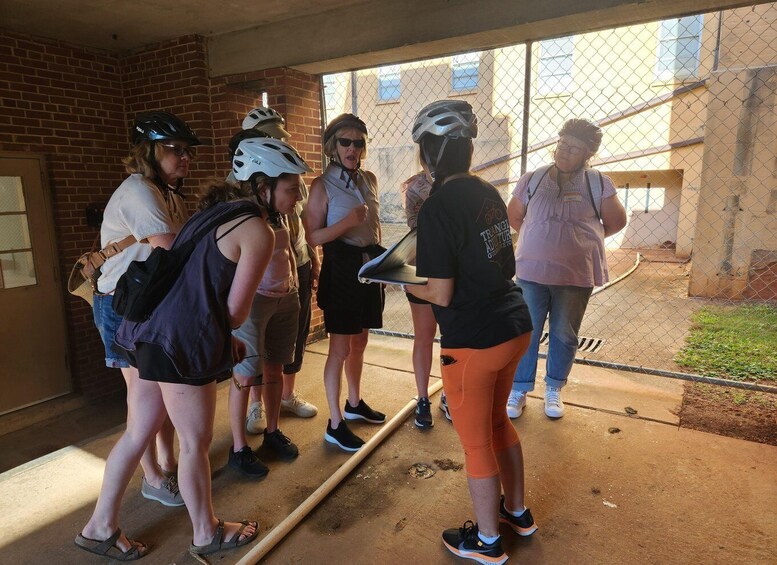 A group of people wearing helmets and casual clothing, standing near a fence and a building with a brick wall. 5