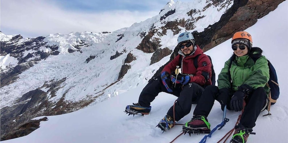 Two people sitting on a snowy mountain slope with rocky terrain and snow-covered peaks in the background. 4